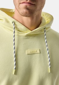 Sudadera con capucha de color amarillo claro y textura acanalada, con un parche de logo en el frente y cordones rayados en azul oscuro y blanco.