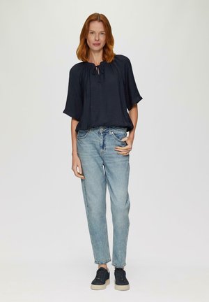 Czarna bluzka z krótkim rękawem z detalem w postaci wiązania przy dekolcie, w połączeniu z jasnoniebieskimi jeansami o prostym kroju i ciemnymi sportowymi butami.