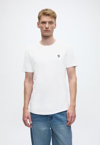 Bijela pamučna majica kratkih rukava s okruglim izrezom i malim crnim logotipom na lijevoj strani prsa. Uparen s svijetloplavim trapericama.