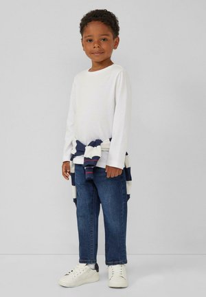 Witte overhemd met lange mouwen, marineblauwe en witte gestreepte trui om de taille gebonden, blauwe denim jeans en witte sneakers met zwarte accenten.