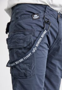 Marine cargobukser med flere lommer, med et stoffmerke som leser "Alpha Industries" og en nøkkelkjede med påskriften "FJERN FØR FLYVNING."