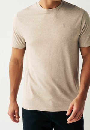 Homme portant un t-shirt beige à manches courtes et col rond, et un pantalon noir, sur un fond clair uni.
