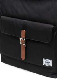 Sac à dos en tissu noir avec un rabat à l'avant, une sangle en cuir marron, une boucle en métal et un écusson blanc avec le logo Herschel dans le coin inférieur.