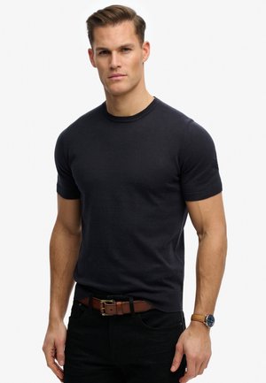 Hombre con el pelo corto que lleva una camisa de punto negra de manga corta, cinturón marrón, pantalones negros y un reloj con correa de cuero marrón.