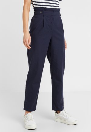 Marineblauwe hoge taille, rechte broek met knoopdetails aan de zijkant, gedragen met witte lage sneakers en een gestreepte ingestopte top.