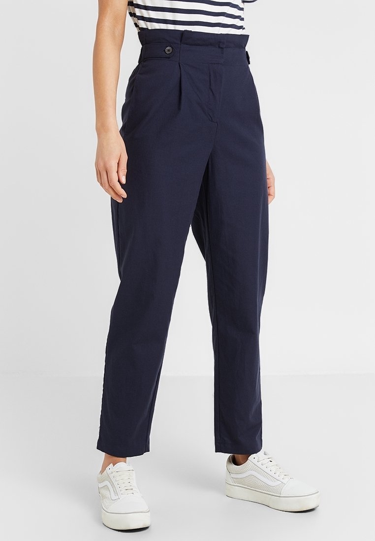 Pantalon bleu marine taille haute à jambe droite avec des boutons sur le côté, porté avec des baskets blanches basses et un haut rayé rentré.