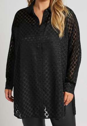 Chemise noire transparente à manches longues avec des pois texturés, boutonnée, portée sur un pantalon noir, vue de la mi-cuisse à la poitrine.