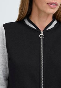 Sort zip-up jakke med grå ærmer, ribbet krave med sort og hvid striber, metallisk lynlås og tekstureret stof.