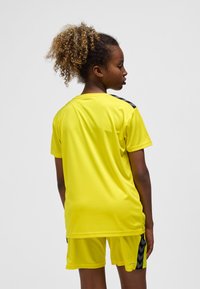 Żółty sportowy zestaw z koszulką i szortami z czarnymi akcentami w kształcie ćwieków na ramionach i bocznych szwach, wykonany z lekkiego, oddychającego materiału.