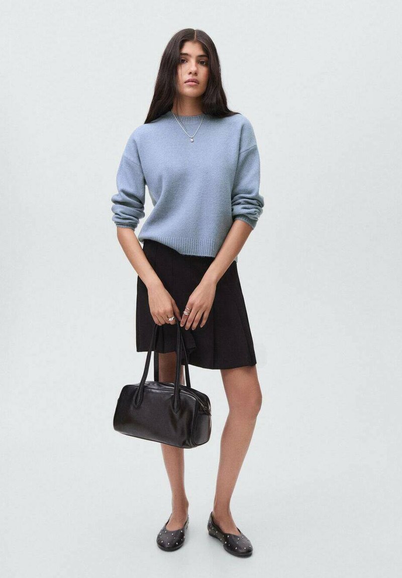Young woman wearing a light blue sweater, black skirt, and black studded flats, holding a black leather handbag with both hands.