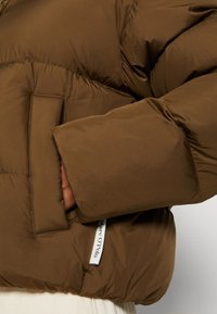 Manche et poche d'une veste matelassée marron avec une main partiellement à l'intérieur, montrant une étiquette en tissu blanche avec le texte "Marc O'Polo".