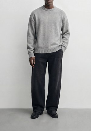 Uomo con maglione in maglia grigio chiaro, pantaloni neri larghi e scarpe nere, in piedi con una mano in tasca su sfondo bianco uniforme.