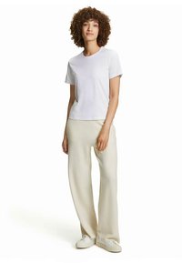 White cotton t-shirt with short sleeves, paired with light beige wide-leg pants. Model stands in casual sneakers, with curly hair.