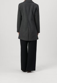 Grå skräddarsydd kappa med en slagkrage, enkel sprund bak, i kombination med löst sittande svarta byxor. Enslig struktur, inga synliga mönster.