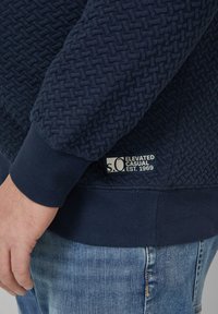 Granatowa bluza z teksturowanym wzorem, ściągaczami na rękawach i naszywką z logo. Materiał ma miękką, tkankową strukturę i delikatny połysk.