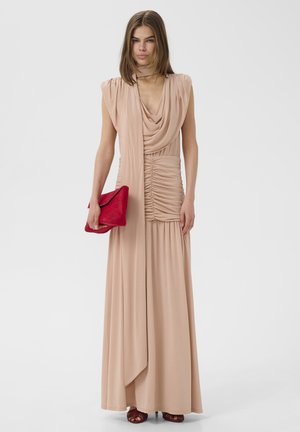 Moteris vilkinti rankovių neturinčią rausvos smėlio spalvos su raukiniais maxi suknelę, laikanti raudoną delninę, stovinti priešais šviesų, vientisą foną.