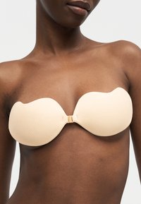 Γυναίκα που φοράει μια ανοιχτόχρωμη, μπεζ αυτοκόλλητη σουτιέν χωρίς έλασμα με υφασμένα καπάκια και κούμπωμα μπροστά πάνω σε σκούρο δέρμα με απλό φόντο.