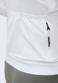 Λευκό υφασμάτινο μπουφάν με ελαστική μέση και κάθετρη τσέπη με φερμουάρ στο πλάι, φορεμένο πάνω από γκρι παντελόνι.