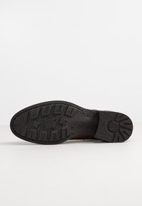 Semelle d’une botte marron foncé présentant une bande de roulement en caoutchouc texturé avec des crampons rectangulaires et un motif circulaire au talon sur fond blanc.