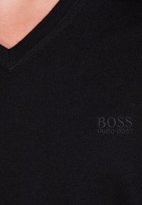 Svart V-ringad tröja gjord av texturerad stickad tyg. Har en subtil präglad logotyp längst ner till vänster. Enkel och ren design.