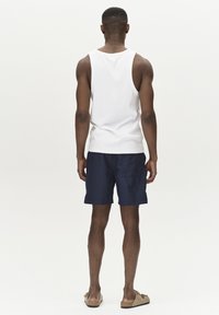 Weißes ärmelloses Tanktop, marineblaue Shorts und beige Sandalen. Das Model ist von hinten zu sehen, was die Farben der Kleidung und das lässige Design betont.