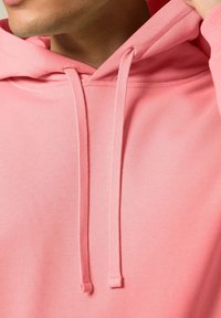 Felpa con cappuccio rosa dal texture morbido, caratterizzata da una tasca a marsupio e cordini regolabili sul cappuccio. La vista ravvicinata mette in evidenza i dettagli del tessuto.