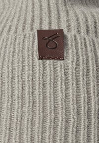Lys grå strikket genser med vertikal ribbestruktur, med et brunt lærpatch med et preget logo-detalj.