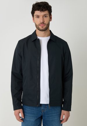 Mężczyzna o kręconych włosach, ubrany w czarną kurtkę na zamek, pod którą ma białą koszulę oraz niebieskie jeansy, stojący na jednolitym jasnym tle.