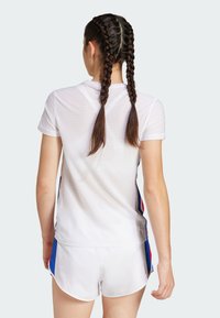 Camisa atlética blanca de manga corta, con textura de malla, acentos laterales azules y rojos; combinada con pantalones cortos blancos, ambos diseñados para deportes.