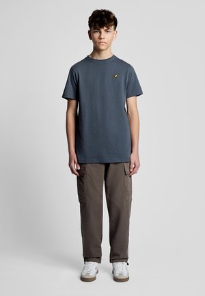 Teenager-Junge steht frontal, trägt ein dunkelgraues T-Shirt mit kurzen Ärmeln, braune Cargohosen und weiße Sneakers vor einem schlichten Hintergrund.