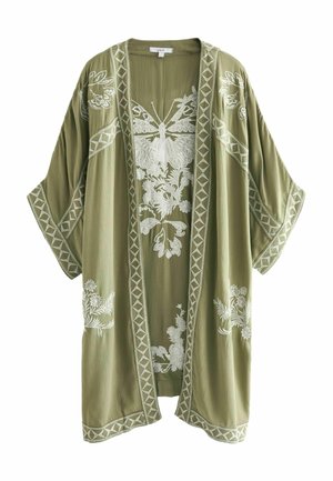 Femme portant un kimono vert clair brodé, un crop top blanc, un short en jean et des sandales beiges, debout contre un mur beige près d'un banc en bois.