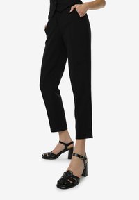 Pantaloni neri su misura con vestibilità slim e tasche laterali, abbinati a sandali con tacco a blocco e cintura alla caviglia, sempre neri.