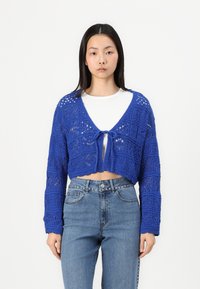 Cropped blue knitted cardigan with openwork pattern, tied at the front. Paired with high-waisted light blue jeans, showcasing a casual look.