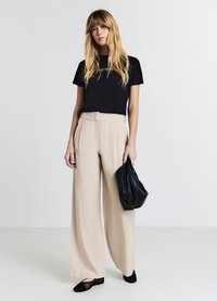 Maglietta nera aderente, pantaloni beige a vita alta e gamba larga, che tiene una borsa nera con texture, con scarpe nere slip-on. Design semplice.