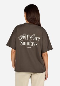 Hnedé oversized tričko s krátkymi rukávmi, na zadnej strane s bielym nápisom "Self Care Sundays." Logo značky pod nápisom.