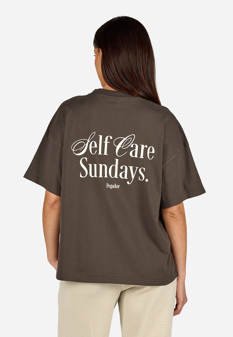 Hnedé oversized tričko s krátkymi rukávmi, na zadnej strane s bielym nápisom "Self Care Sundays." Logo značky pod nápisom.