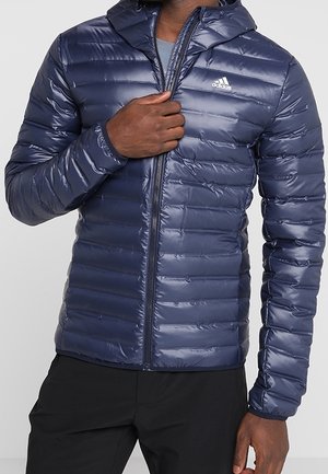Homme portant une veste matelassée Adidas bleu marine à capuche, partiellement zippée, avec un pantalon noir sur un fond clair uni.