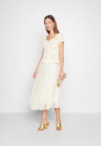 Top din dantelă în alb murdar, cu o textură florală, asociat cu o fustă de tull ivorie, fluidă și plisată. Accesorizat cu mule cu toc aurii și o geantă de umăr.