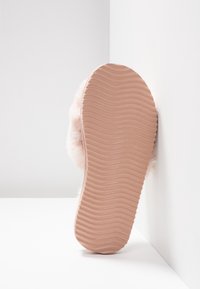 Semelle d'une pantoufle rose avec une semelle ondulée et texturée appuyée contre des murs blancs et un sol, avec des accents de fourrure douce visibles.