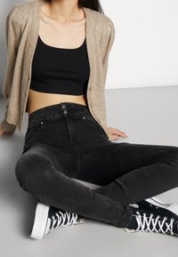 Cárdigan de punto beige, top corto negro, jeans negros de talle alto con cierre de botón y zapatillas negras con suelas y cordones blancos.