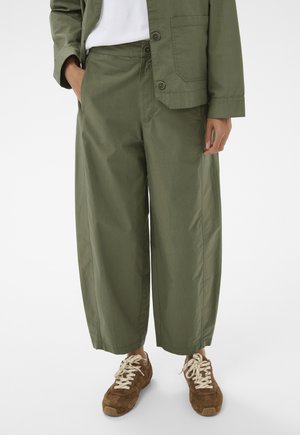 Person trägt olivgrüne, gekürzte Hose mit weitem Bein, passende olivgrüne Jacke, weißes Hemd und braune Wildledersneaker mit weißen Schnürsenkeln.