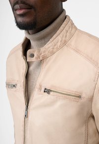 Mand iført en beige læderjakke med lynlåslommer på brystet over en lys brun turtleneck striktrøje, delvist ansigt synligt.