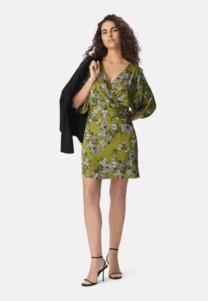 Abito verde floreale con scollo a V, maniche ampie, vita cinchata e lunghezza corta; abbinato a sandali neri con tacco alto e un blazer.