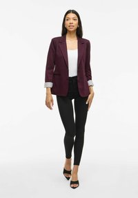Femeie mergând înainte, îmbrăcată cu un sacou burgund, un top alb, pantaloni negri skinny și sandale negre cu vârf deschis pe un fundal alb simplu.