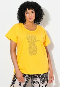 Camiseta de manga corta amarilla con cuello redondo, que presenta un diseño de piña brillante en el centro. Tejido suave y corte relajado.
