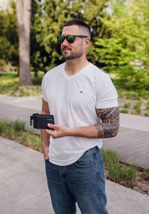 Black wallet with a zipper and clip held by a man in a white V-neck shirt and blue jeans. Sunglasses reflect green trees.