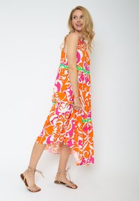 Farbenfrohes ärmelloses Kleid mit einem orange-rosa-weißen Blumenmuster, mit einem Rüschensaum und grünen Akzenten. Kombiniert mit hellbraunen Sandalen.