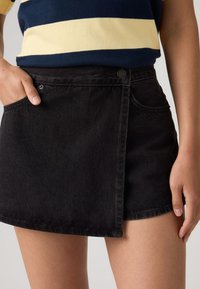 Άτομο που φοράει μαύρη ασύμμετρη φούστα από τζιν με τσέπες, συνδυασμένη με αμάνικο τοπ με οριζόντιες ρίγες σε ναυτικό μπλε και κρεμ χρώ μα.