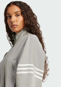 Chaqueta gris con cremallera que presenta un cuello alto y rayas blancas acanaladas a lo largo de las mangas. La tela parece suave y tiene un corte relajado.