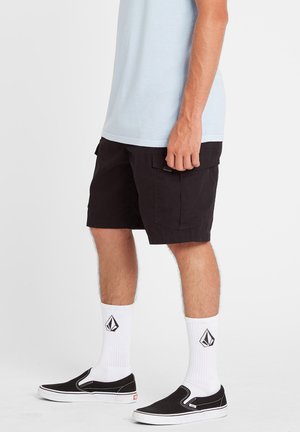 Shorts cargo noirs avec poches, portés avec des chaussettes blanches à logo et des chaussures noires à enfiler, associés à un t-shirt bleu clair.